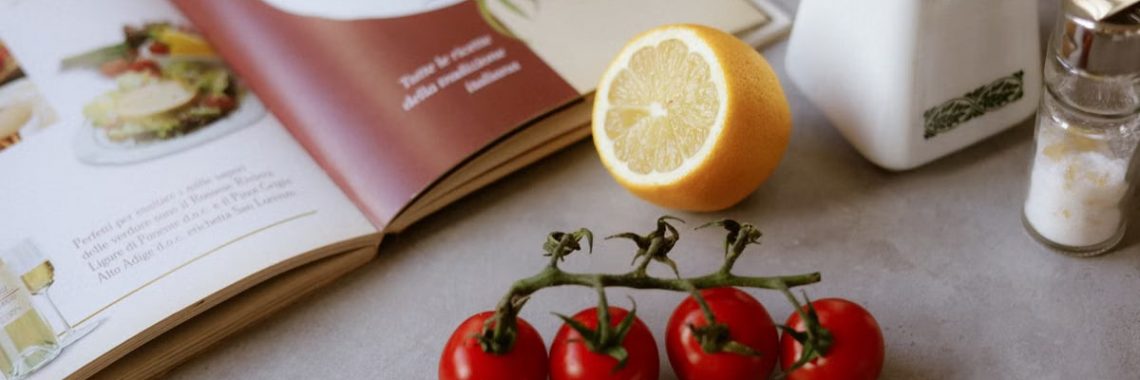 Flat lay of cooking ingredients including tomatoes and a lemon with a cookbook.